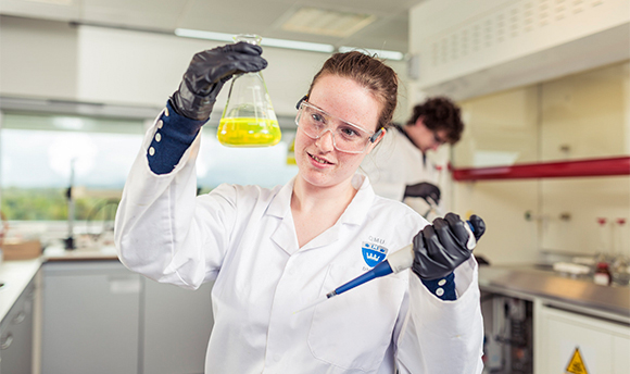 A 69ý student wearing PPE and holding up a beaker of yellow liquid